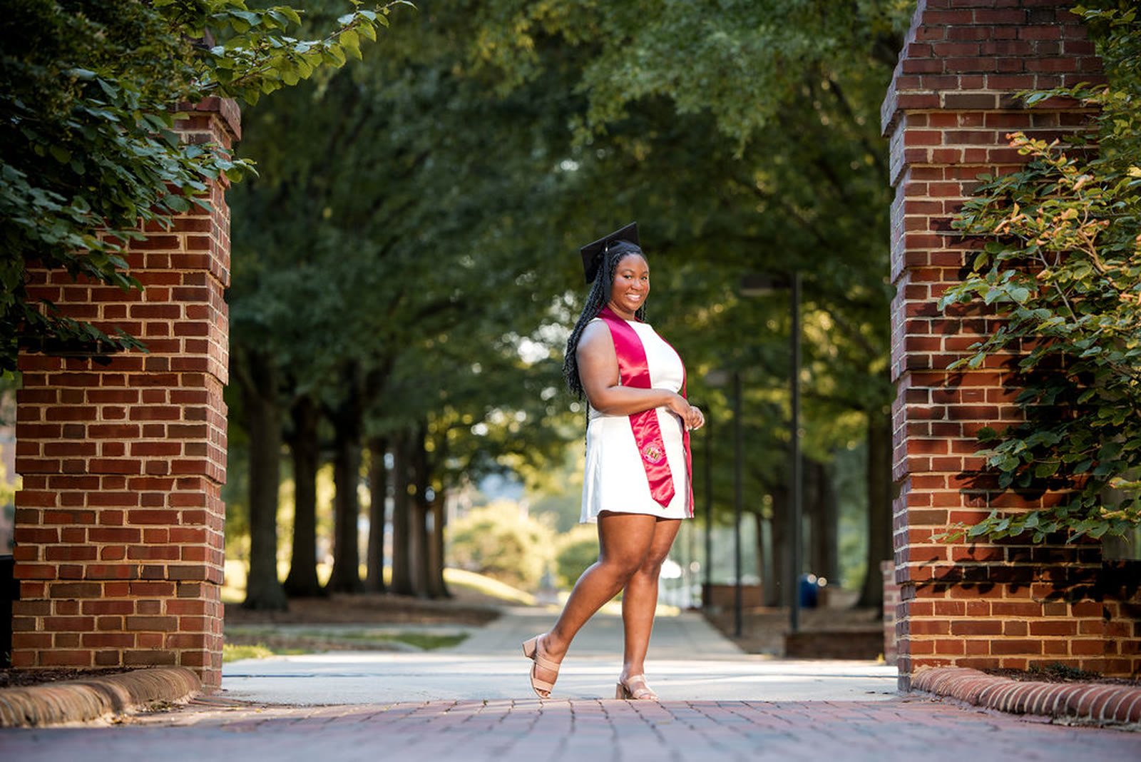Graduate walking a tree lined campus path framed by brick pillars